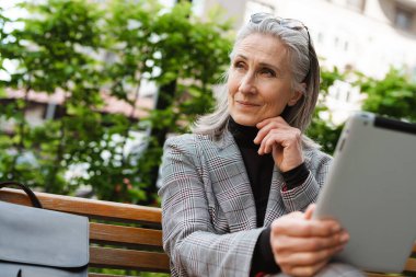 Açık havada bankta otururken tablet bilgisayar kullanan gri beyaz bir kadın.