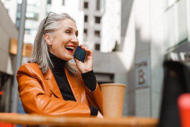 Kafede otururken cep telefonuyla konuşan son sınıf öğrencisi gri beyaz bir kadın.