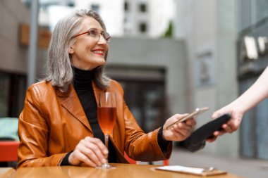 Dışarıda kafede şarap içerken cep telefonuyla ödeme yapan yaşlı gri bir kadın.