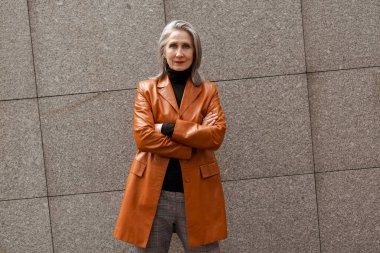 Dışarıda dikilirken kameraya bakan deri ceketli gri yaşlı kadın.