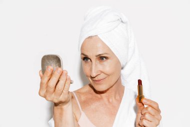 Beyaz duvardan izole edilmiş ruj sürerken havluyla gülümseyen yaşlı kadın.