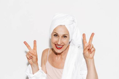 Beyaz duvarda izole edilmiş barış işareti yaparken havluyla gülümseyen yaşlı kadın.