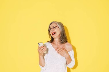 Sarı duvarda izole edilmiş cep telefonu kullanan gözlüklü, gülen olgun bir kadın.