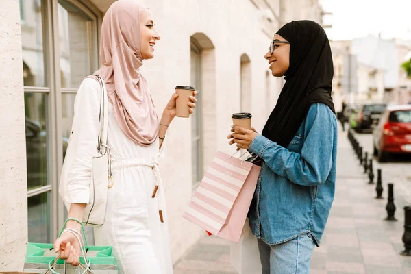 İki orta yaşlı, başörtüsü takmış gülümseyen İslamcı kadın ellerinde kahve fincanlarıyla ellerinde poşetlerle dışarıda yürüyorlar.