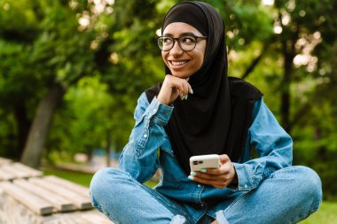Gülümseyen orta yaşlı Müslüman kadın bir bankta oturmuş cep telefonu kullanıyor.