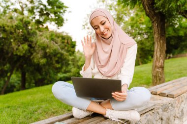 Tesettürlü genç müslüman kadın dizüstü bilgisayarla bankta oturuyor.