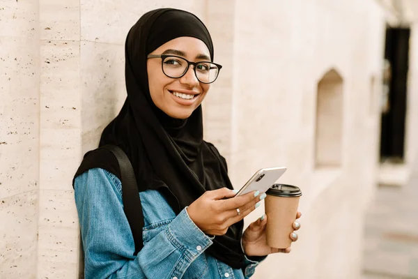 一名穿着头巾 面带微笑的年轻穆斯林妇女站在街上 用手机喝咖啡 — 图库照片