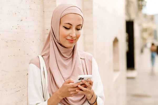 一名穿着头巾 面带微笑的年轻穆斯林妇女用手机站在街上 — 图库照片
