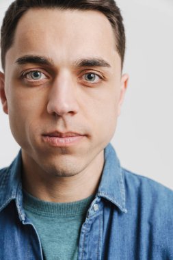 Genç esmer beyaz adam poz veriyor ve beyaz arka planda izole edilmiş kameraya bakıyor.