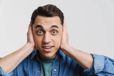 Genç heyecanlı adam kulaklarını kapatıyor ve beyaz arka planda izole edilmiş kameraya bakıyor.