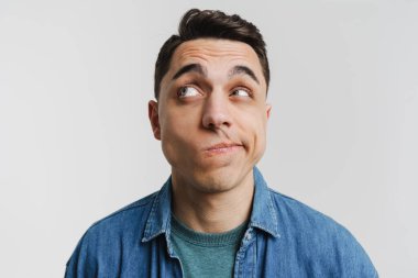 Genç, esmer, kot gömlekli, suratını buruşturan ve beyaz arka planda soyutlanmış bir şekilde bakan adam.