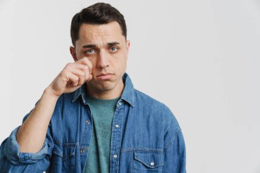 Genç esmer adam beyaz arka planda izole edilmiş kameraya ağlarken gözyaşlarını siliyor.