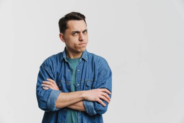 Genç esmer adam beyaz arka planda kollarını kavuştururken kaşlarını çatıyor.