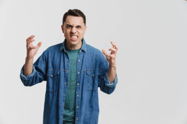 Genç esmer adam beyaz arka planda izole edilmiş kot gömlekli, el kol hareketi yapan ve kameraya somurtan.