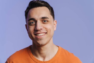 Genç esmer adam gülümsüyor ve mor arka planda izole edilmiş kameraya bakıyor.