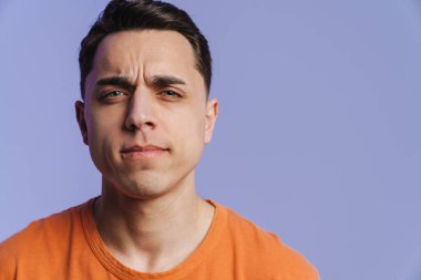 Genç esmer adam somurtuyor ve mor arka planda izole edilmiş kameraya bakıyor.