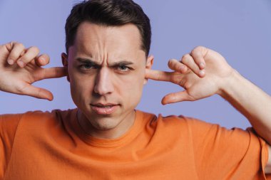 Genç esmer adam kaşlarını çatıyor ve kulaklarını tıkıyor. Mor arka plan için izole edilmiş.