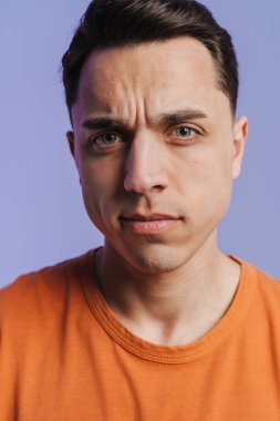 Genç esmer beyaz adam somurtuyor ve mor arka planda izole edilmiş kameraya bakıyor.