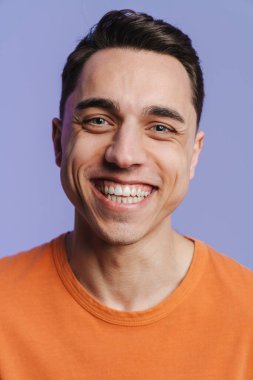 Genç esmer beyaz adam gülümsüyor ve mor arka planda izole edilmiş kameraya bakıyor.