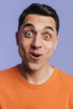 Genç esmer beyaz adam mor arka planda izole edilmiş kameraya şaşırdığını ifade ediyor.