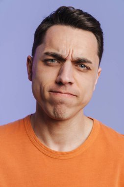 Genç esmer beyaz adam somurtuyor ve mor arka planda izole edilmiş kameraya bakıyor.