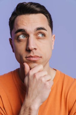 Genç esmer beyaz adam düşünüyor ve mor arka planda izole edilmiş bir şekilde yan tarafa bakıyor.