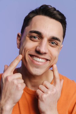 Genç esmer beyaz adam, mor arka planda izole edilmiş gülüşünü işaret ediyor.