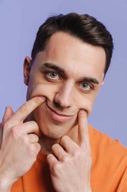 Genç esmer beyaz adam gülümsüyor, parmakları mor arkaplanda soyutlanmış.