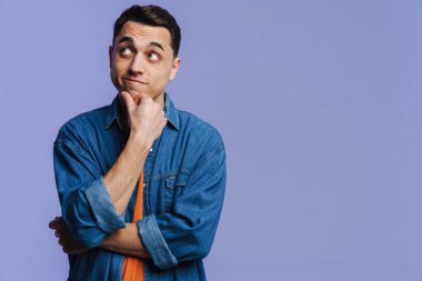 Kot pantolon giymiş genç esmer adam mor arka planda izole edilmiş bir şekilde düşünüyor ve yan bakıyor.