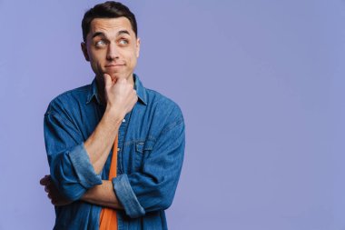 Genç esmer beyaz adam düşünüyor ve mor arka planda izole edilmiş bir şekilde yan tarafa bakıyor.