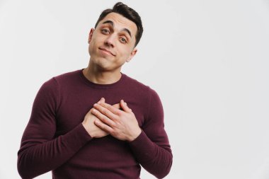 Genç esmer adam göğsünde el ele tutuşuyor ve beyaz arka planda izole edilmiş kameraya bakıyor.