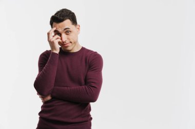 Genç esmer adam yüzünü kapatıyor ve beyaz arka planda izole edilmiş kameraya bakıyor.