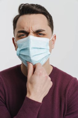 Yüzü maskeli genç esmer adam beyaz arka planda izole edilmiş kameraya bakıyor.