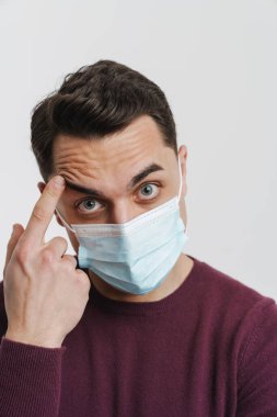 Yüzünde maske olan genç esmer adam beyaz arka planda izole edilmiş alnını işaret ediyor.