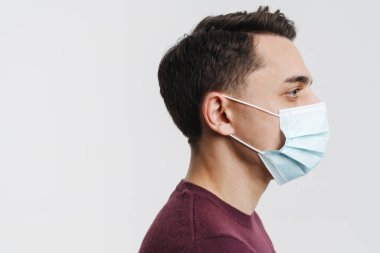 Yüz maskeli genç esmer adam profilde poz veriyor ve beyaz arka planda soyutlanmış bir şekilde ileriye bakıyor.
