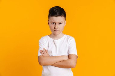 Sarı arka plan kolları katlanmış tişörtlü, asık suratlı çocuk.