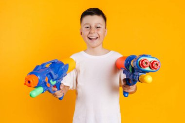 Sarı arka planda duran ve su tabancasıyla oynayan mutlu çocuk tişörtlü çocuk.