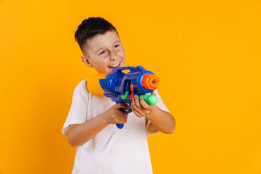 Sarı arka planda duran ve su tabancasıyla oynayan mutlu çocuk tişörtlü çocuk.