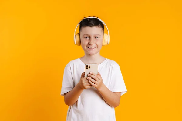 黄色の背景に隔離されたヘッドフォンや携帯電話を使用している間 ブルネットの白い男の子笑顔 — ストック写真
