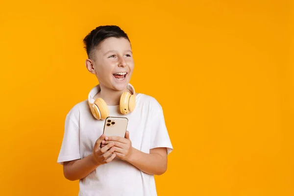 黄色の背景に隔離された携帯電話を使用している間に笑ってヘッドフォンを持つブルネットの白い少年 — ストック写真
