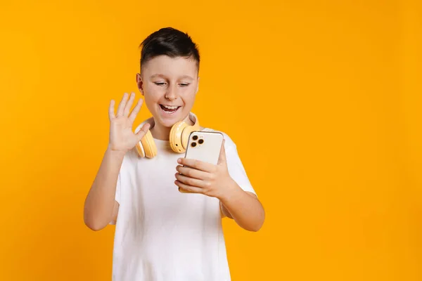 黄色の背景に隔離された携帯電話を使用している間 ヘッドフォンジェスチャー付きブルネットの白い少年 — ストック写真