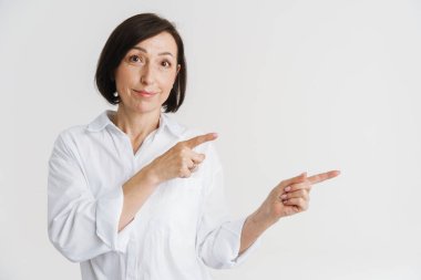 Avrupalı olgun bir kadın gülümsüyor ve parmaklarını beyaz duvarın üzerinde soyutlanmış bir şekilde gösteriyor