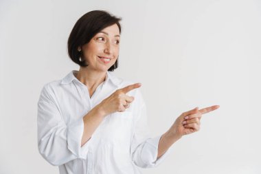 Avrupalı olgun bir kadın gülümsüyor ve parmaklarını beyaz duvarın üzerinde soyutlanmış bir şekilde gösteriyor