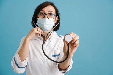 Avrupalı maske takan, maske takan, steteskopla mavi duvarın üzerinde duran olgun bir doktor.