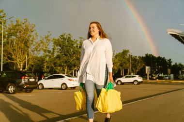 Dışarıdaki park yerinde alışveriş torbalarıyla yürürken gülümseyen genç beyaz kadın.