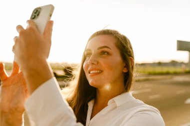 Genç beyaz kadın dışarıda park yerinde cep telefonuyla selfie çekiyor.