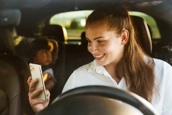 車の中で息子と座っている間 白い女性は笑顔と携帯電話を使用して — ストック写真