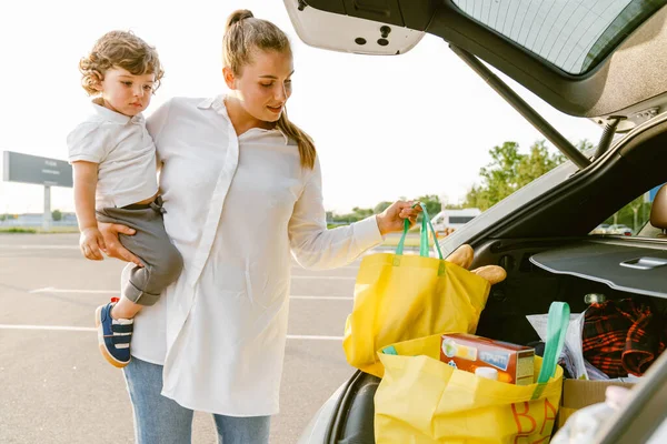駐車場でトランクから買い物袋を取得しながら 白人女性は彼女の息子を保持 — ストック写真