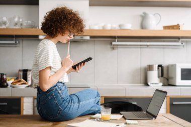 Ev mutfağında dizüstü bilgisayarla çalışırken cep telefonu kullanan ve elinde cep telefonu olan genç bir kız öğrenci.
