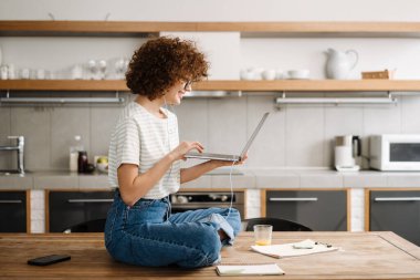 Avrupa 'dan gülümseyen kadın mutfaktaki masada otururken dizüstü bilgisayar ve kulaklık kullanıyor.
